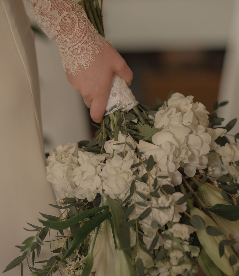 Bride holding a lush white hydrangea and greenery wedding bouquet, wearing a lace sleeve gown.