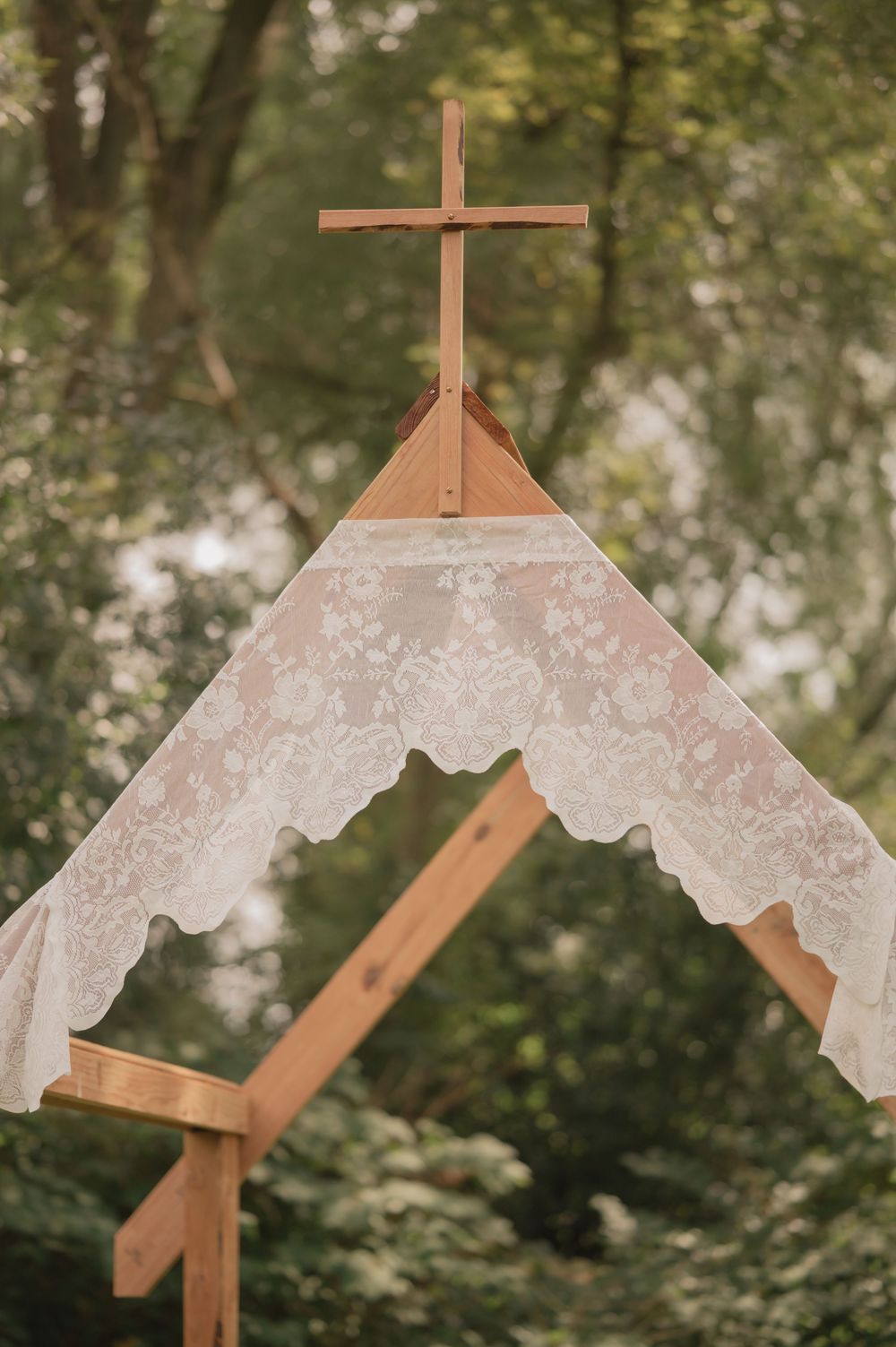 Rustic wooden wedding arch with lace draping and cross in a lush green forest setting.