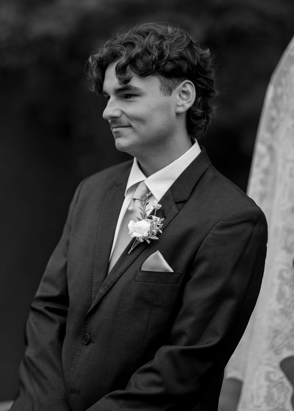 Young groom in dark suit with boutonniere smiling at wedding ceremony, black and white portrait photo.