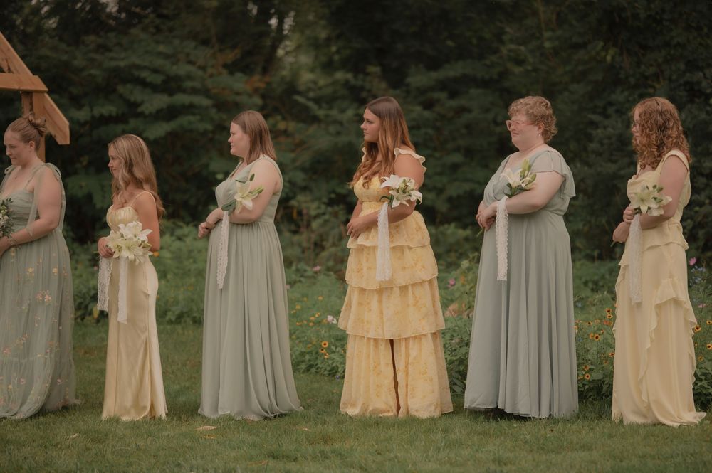 Six bridesmaids in sage green and yellow gowns holding bouquets outdoors at a wedding ceremony.