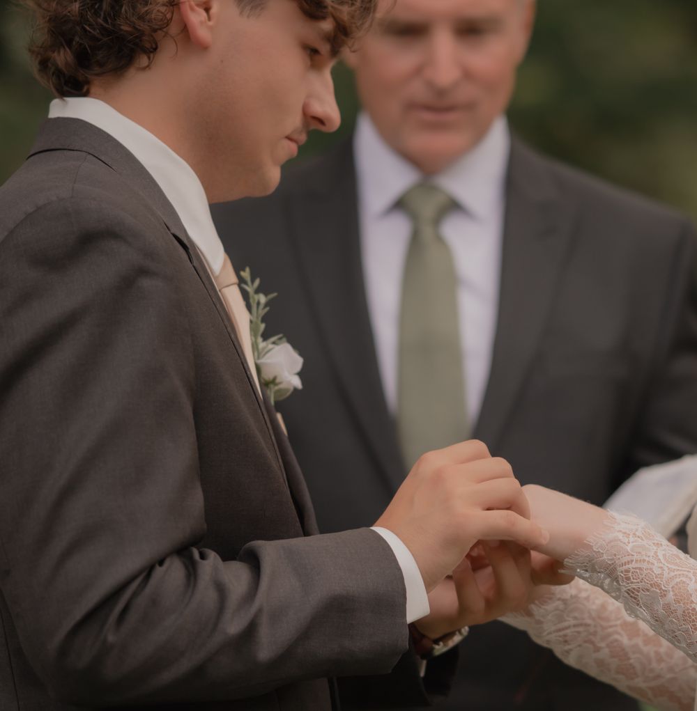 Groom placing wedding ring on bride's finger during outdoor ceremony, officiant standing in background.
