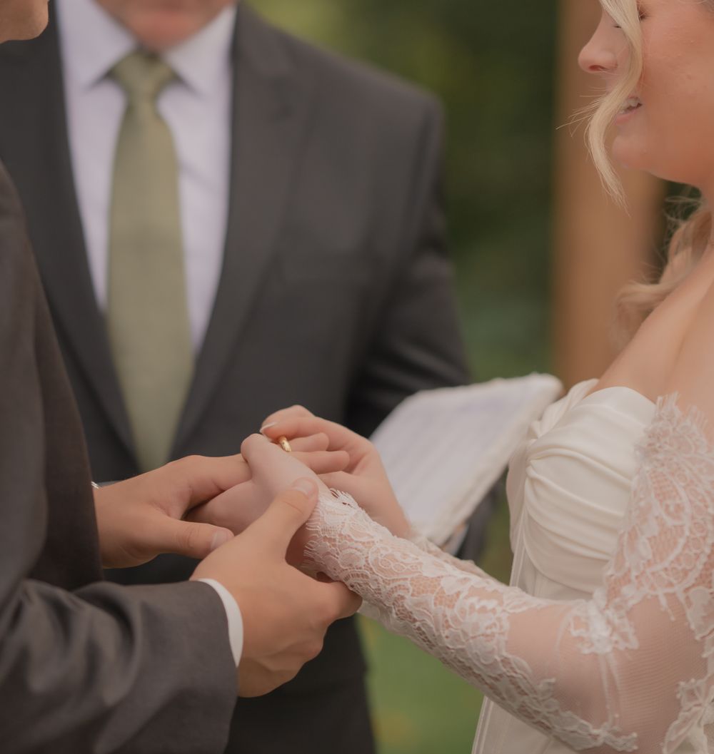A groom places a gold wedding ring on his bride's finger during an outdoor wedding ceremony.