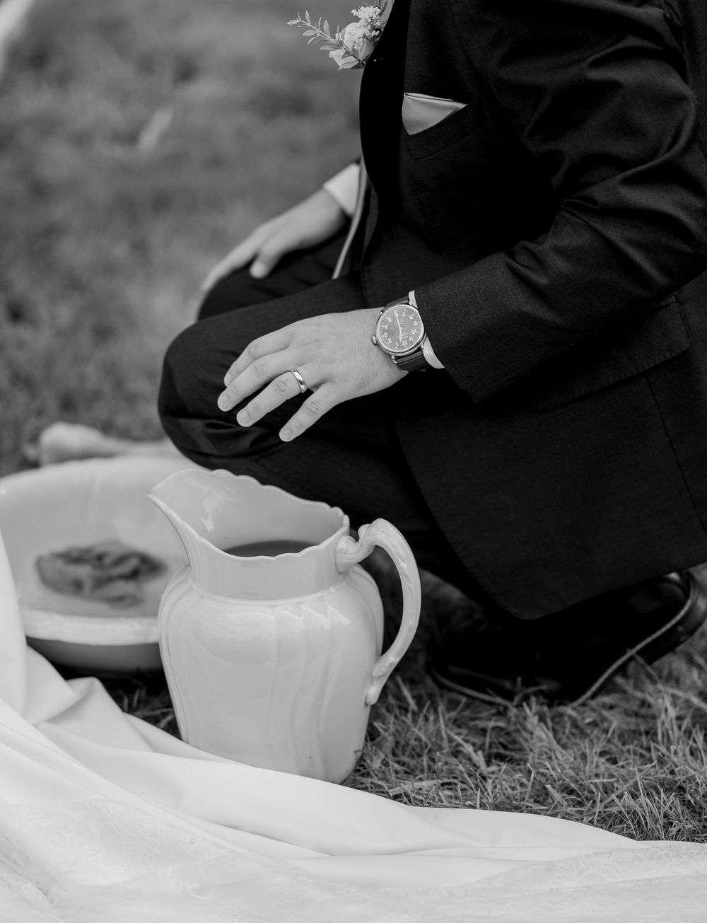 Black and white photo of a groom in a suit kneeling on grass beside a white ceramic pitcher and plate.