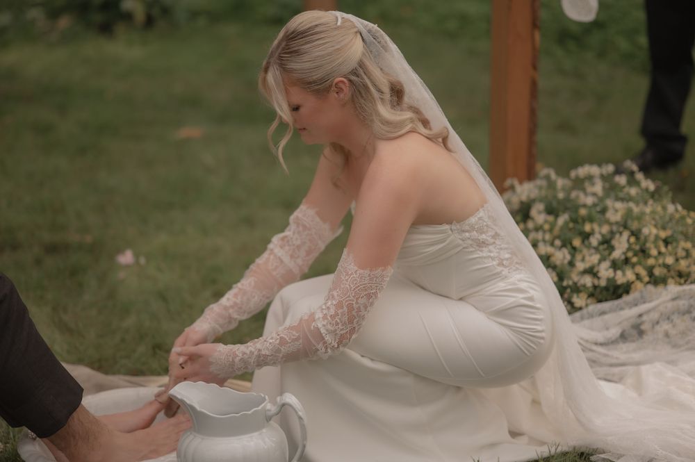 Bride in white gown and lace gloves performing a foot washing ceremony outdoors during a wedding.