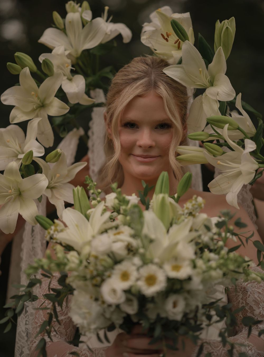 A smiling bride holding a white bouquet surrounded by elegant white lily flowers on her wedding day.