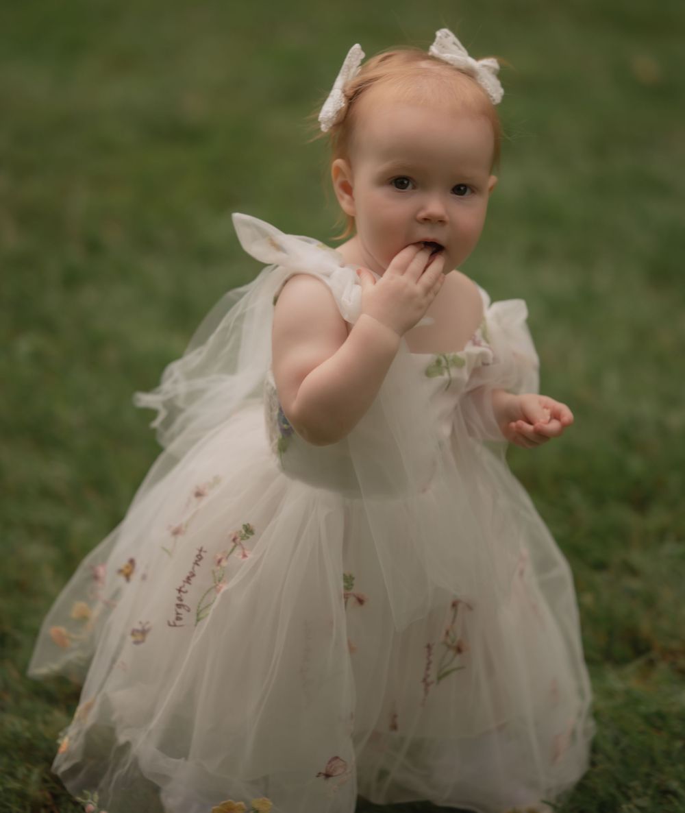 A baby girl in a white floral tulle dress and bow headband plays outdoors on green grass.
