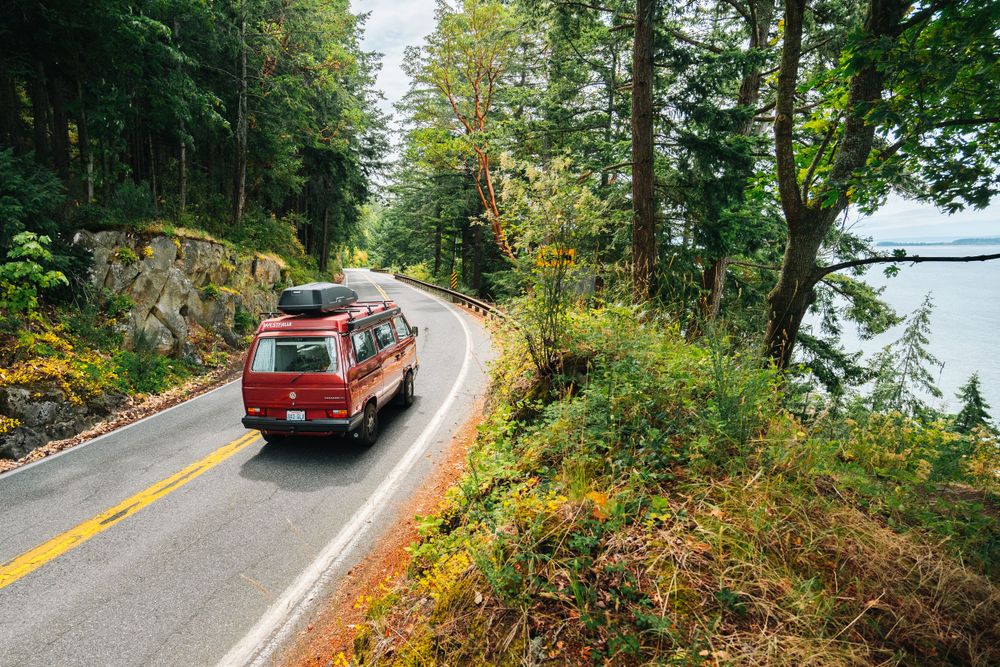 Red vintage van driving along winding coastal road through lush forest during autumn season.
