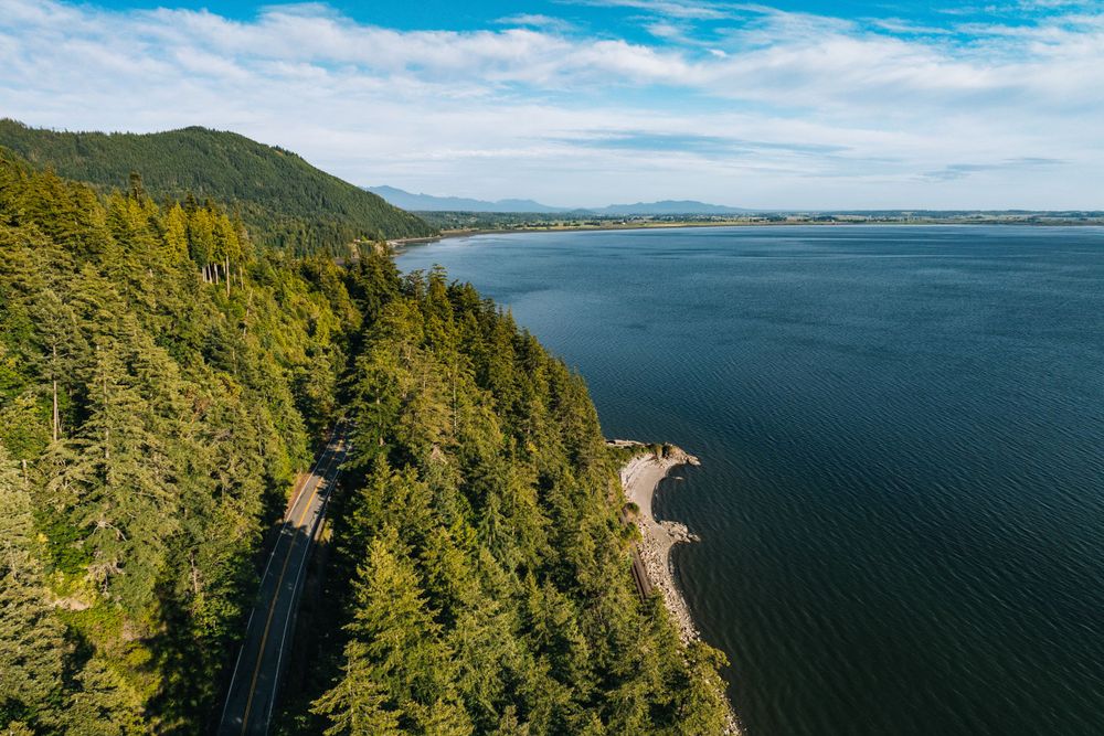 Aerial view of a winding coastal road along evergreen forest and blue waters of the Pacific Northwest.