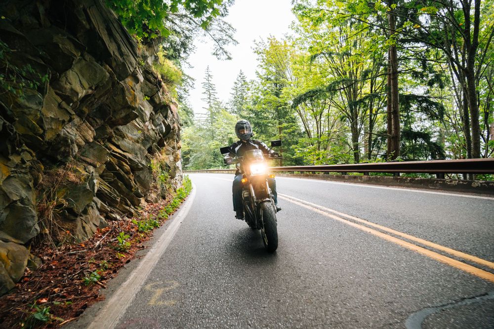 A motorcyclist rides on a winding forest road with rock wall and green trees in the background.