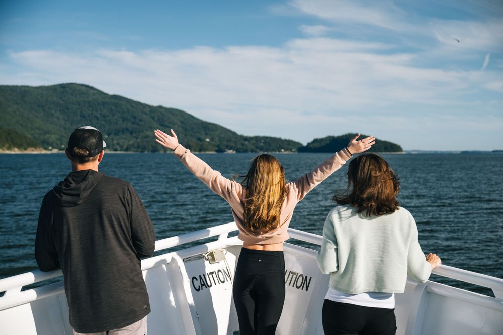 Three people enjoying a boat ride on the water with mountains and blue sky in the background as one extends their arms.