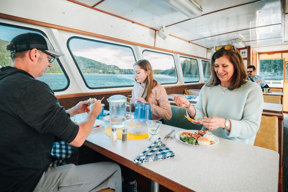 Passengers enjoy a meal and drinks at a booth table aboard a scenic boat cruise.