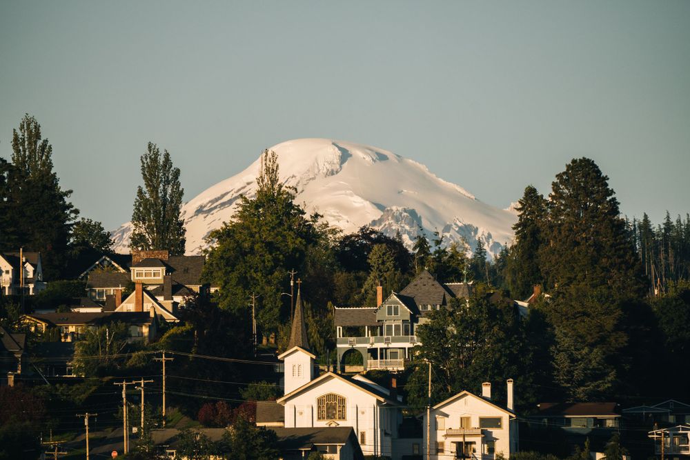 Snow-capped Mount Baker rises above residential homes and evergreen trees in Washington state during golden hour.