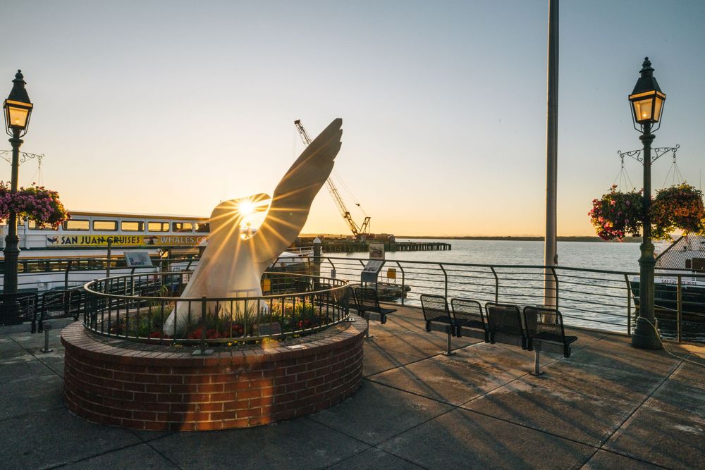A sundial sculpture on a waterfront walkway captures golden hour sunlight with decorative lampposts at sunset.