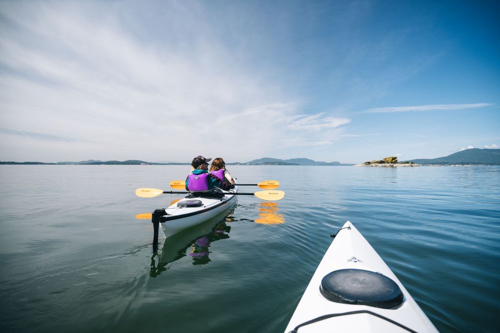 Two people kayaking on calm waters with mountains in the distance under blue skies.