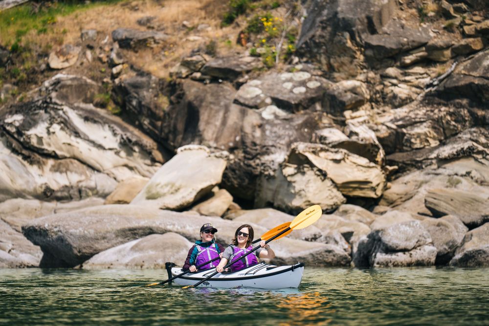 Two kayakers paddle along rocky cliffs on a scenic lake in a double kayak.