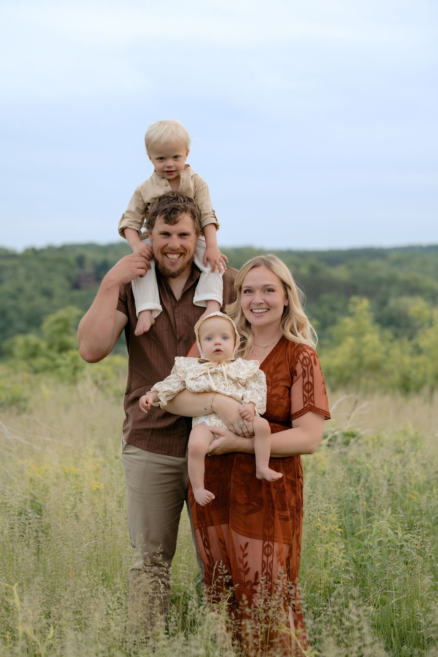 A young mom and dad hold their baby and toddler in a grassy hilly field smiling at the camera for family photos in Hudson, WI