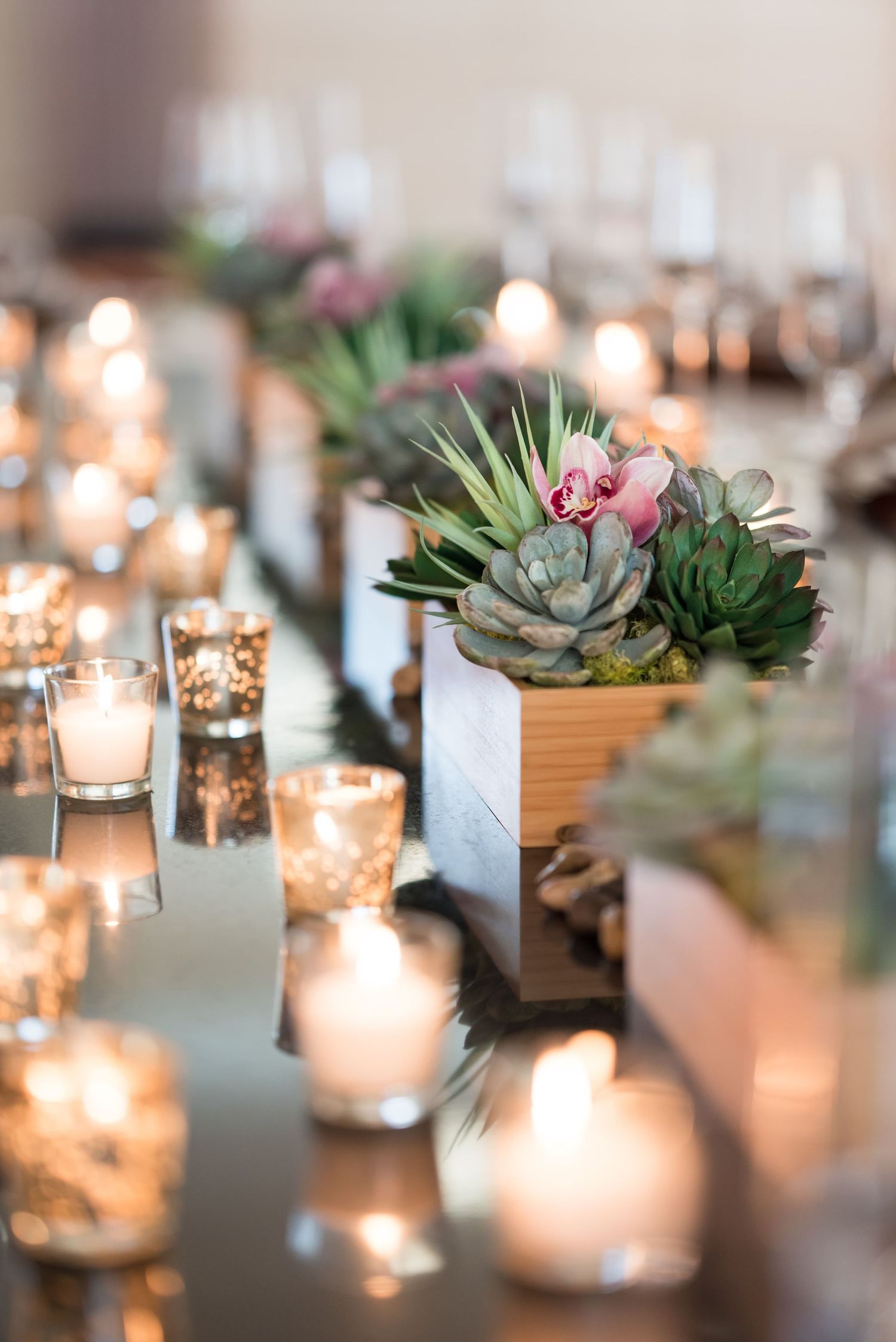 Elegant wedding table centerpiece with succulent arrangements in wooden boxes surrounded by glowing votive candles.