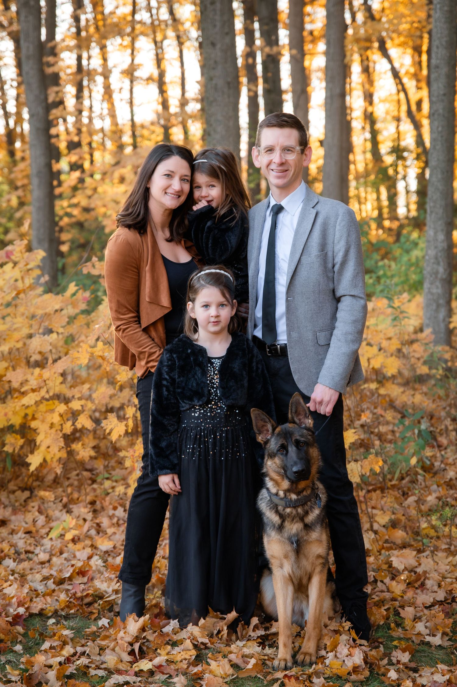 Family of four with a white dog posing outdoors on grass surrounded by autumn trees.