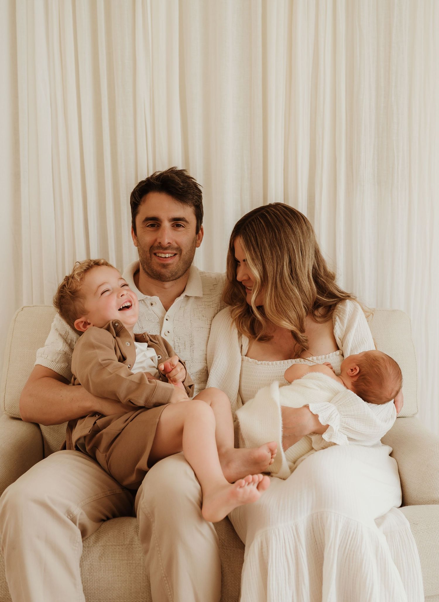 Happy family of four with toddler laughing, dad smiling, and mom holding newborn baby on a cream sofa.