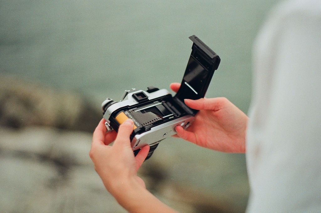 Person holding an open vintage film camera near a rocky shoreline, loading or inspecting the film compartment.