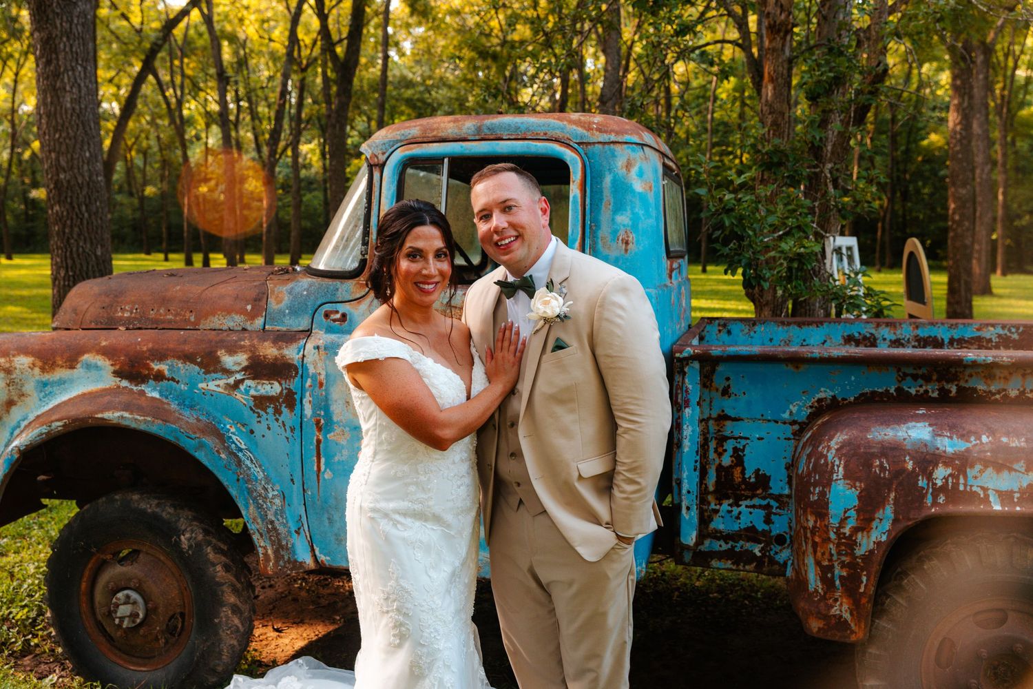 Bride and groom pose before a rustic blue vintage truck outdoors at a wedding venue surrounded by trees.
