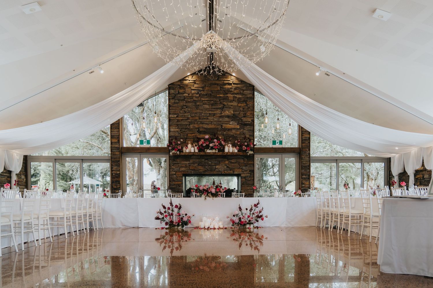 Elegant wedding venue with white draped ceiling, crystal chandelier, stone fireplace, and floral arrangements on a polished floor.