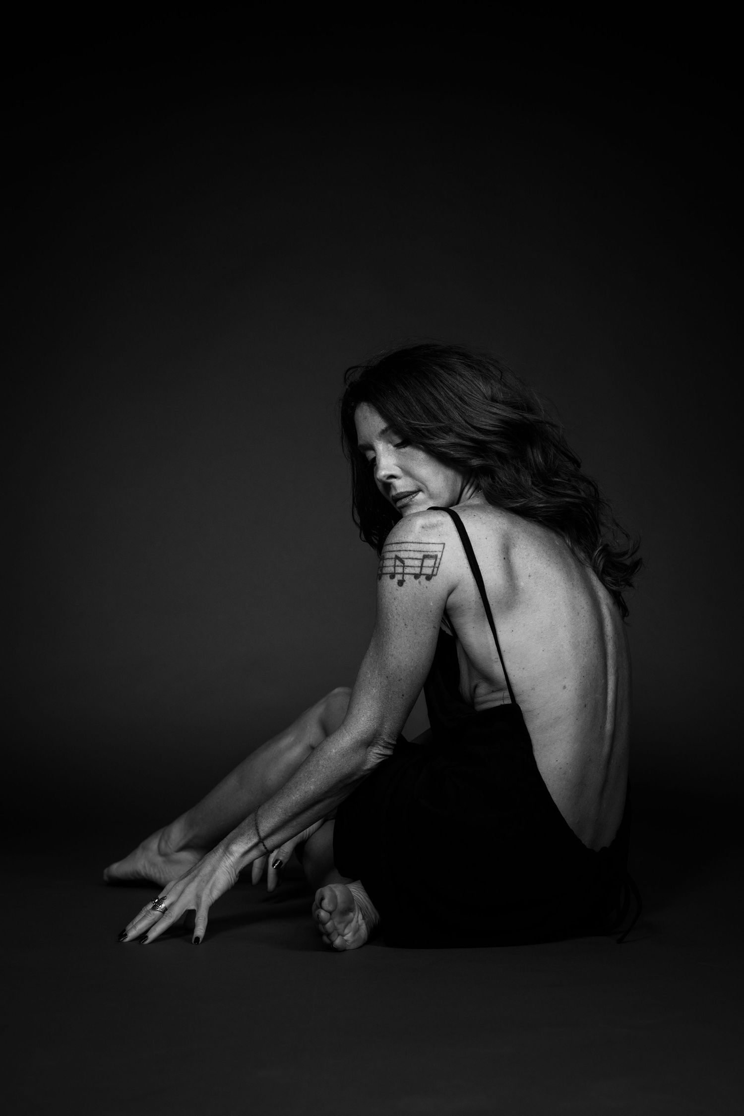 Black and white studio photo of a woman in a backless black dress sitting on the floor.
