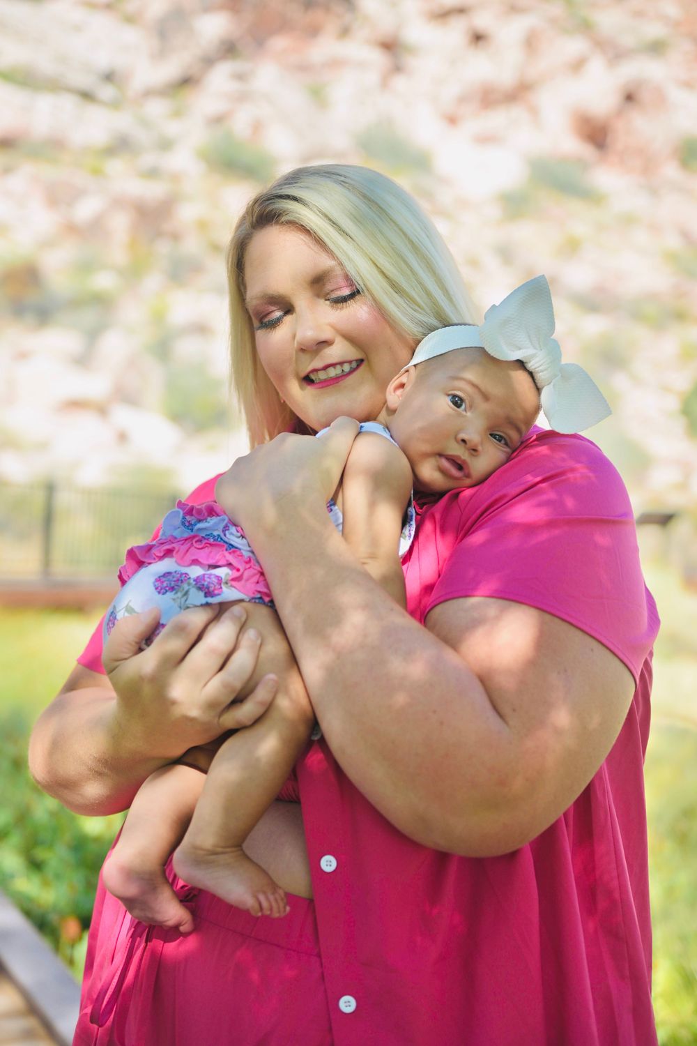 Blonde woman in pink dress tenderly holds two infants outdoors with rocky landscape background.