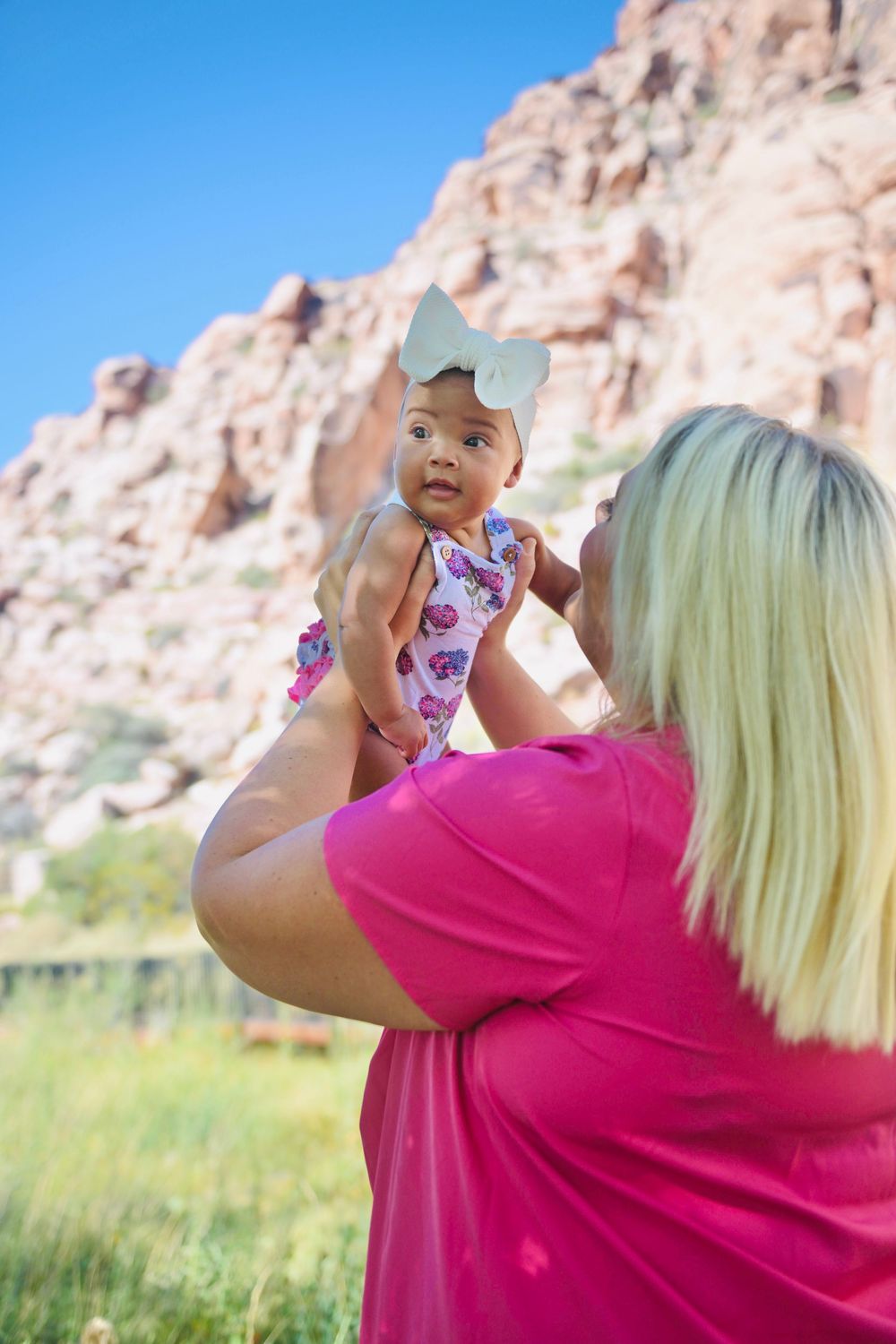 A blonde woman in pink holds a baby girl in a floral outfit with a white bow at Red Rock Canyon.