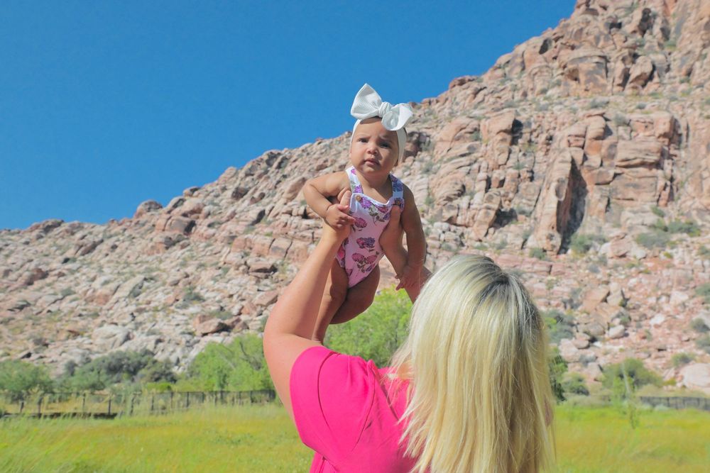 A blonde woman in pink lifts a baby girl with a bow against a rocky desert landscape.