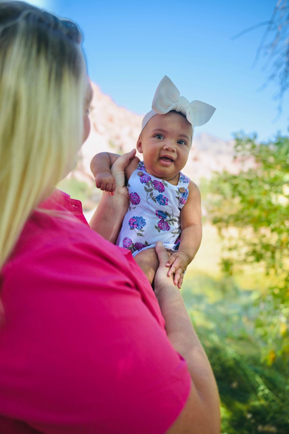 A blonde woman in a pink shirt holds up a smiling baby girl in a floral outfit with a white bow outdoors.