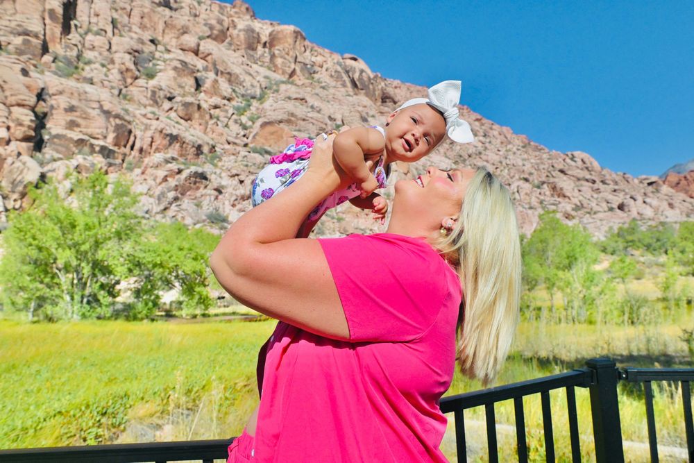 Woman in pink shirt lifting smiling baby girl with bow against scenic red rock canyon and green trees backdrop.