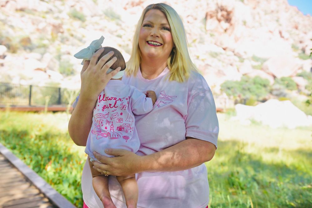 Woman smiling outdoors holding a baby dressed in pink near rocky landscape and green trees.