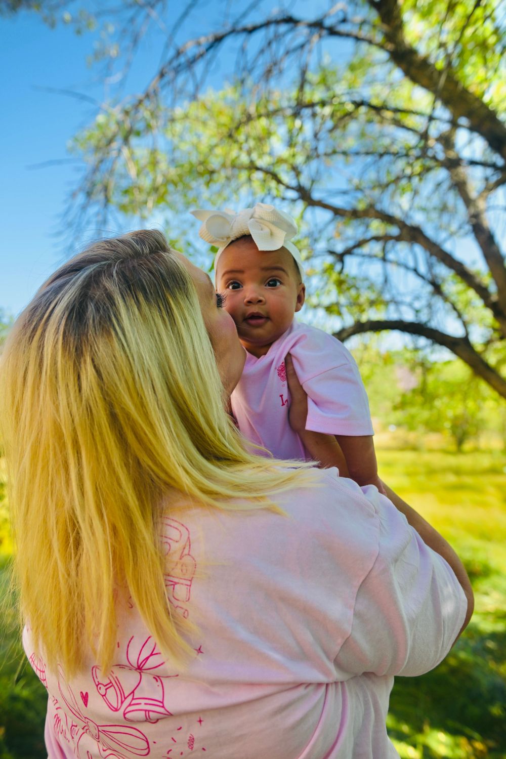 Blonde woman kissing a baby girl in a pink outfit with a white bow outdoors under a tree.