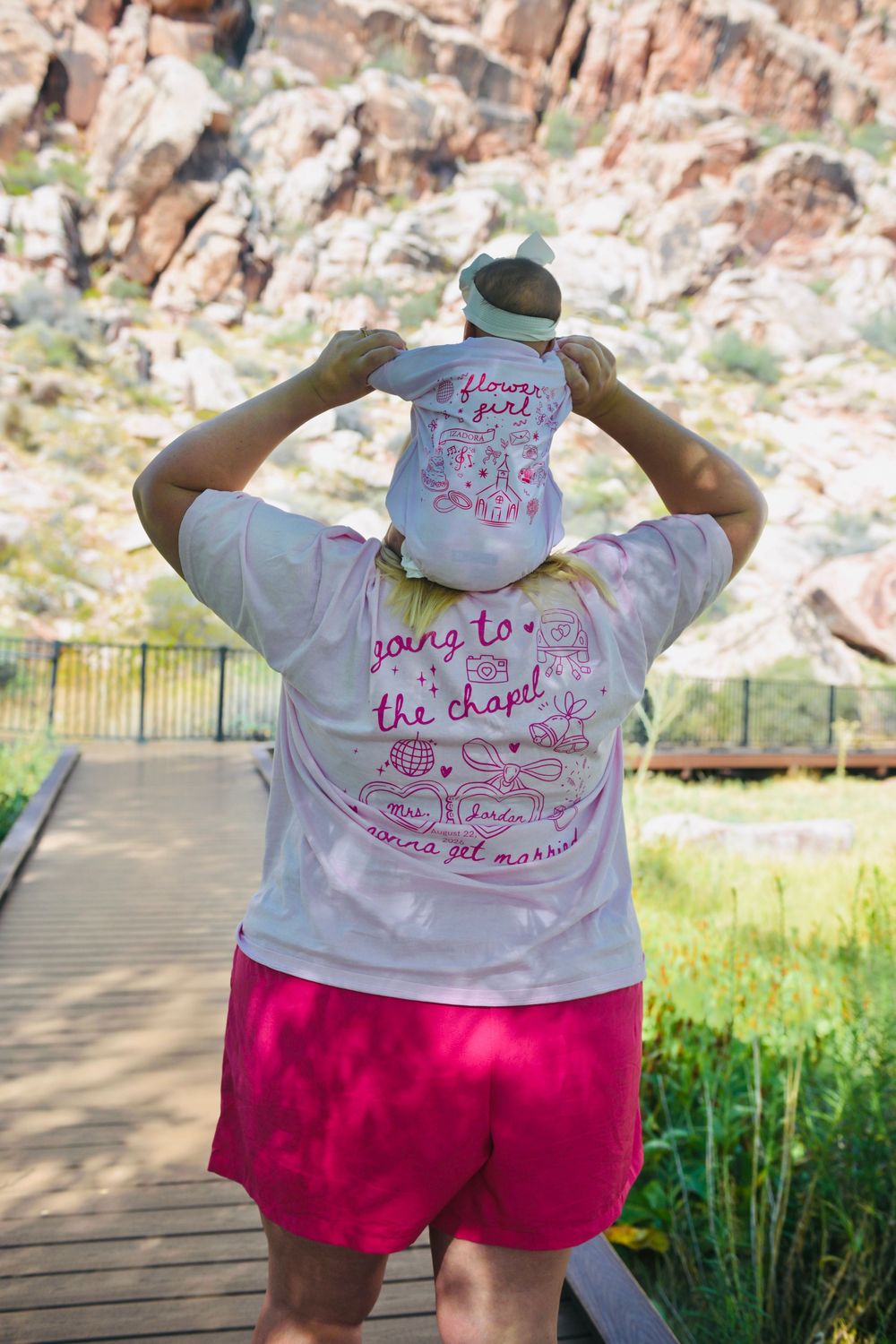 Woman in a pink bridal t-shirt holds a baby on her shoulders at a scenic rocky landscape.