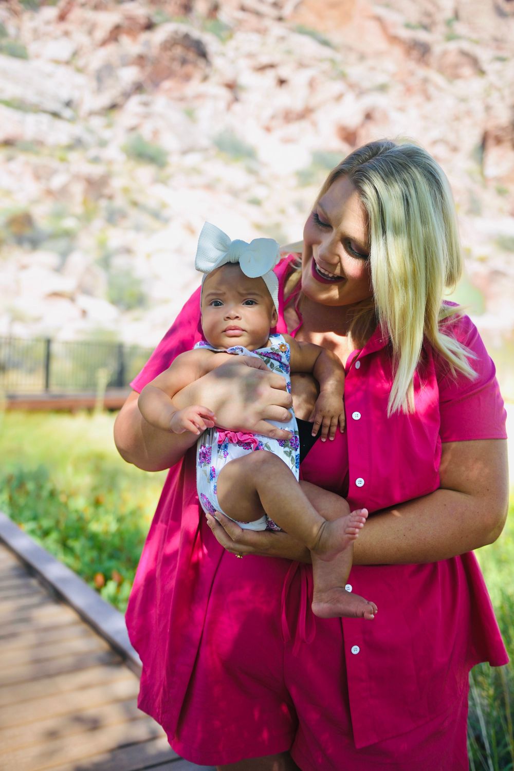 Blonde woman in pink dress holding a baby girl with a bow headband outdoors near red rock formations.