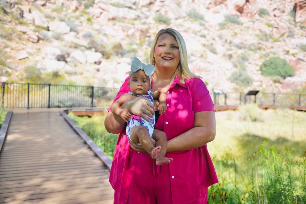 Woman in pink dress holding baby outdoors near rocky landscape and wooden bridge on sunny day.
