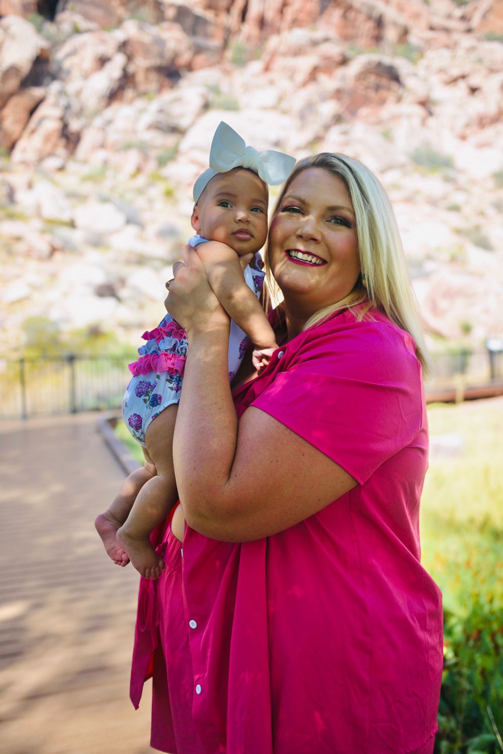 Smiling blonde woman in a pink dress holds a baby girl with a bow headband outdoors.