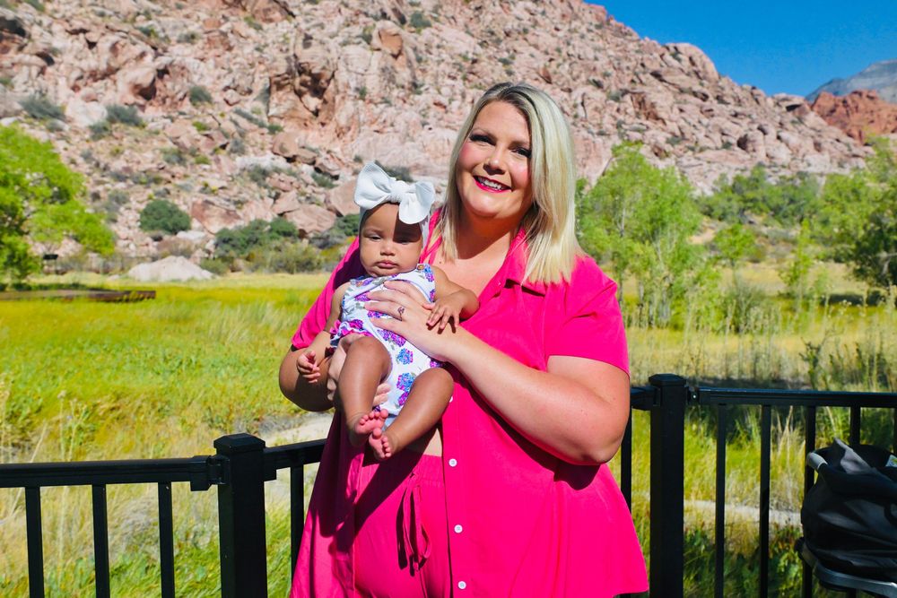 Woman in pink dress holding baby girl outdoors with red rock mountains and green meadow background.