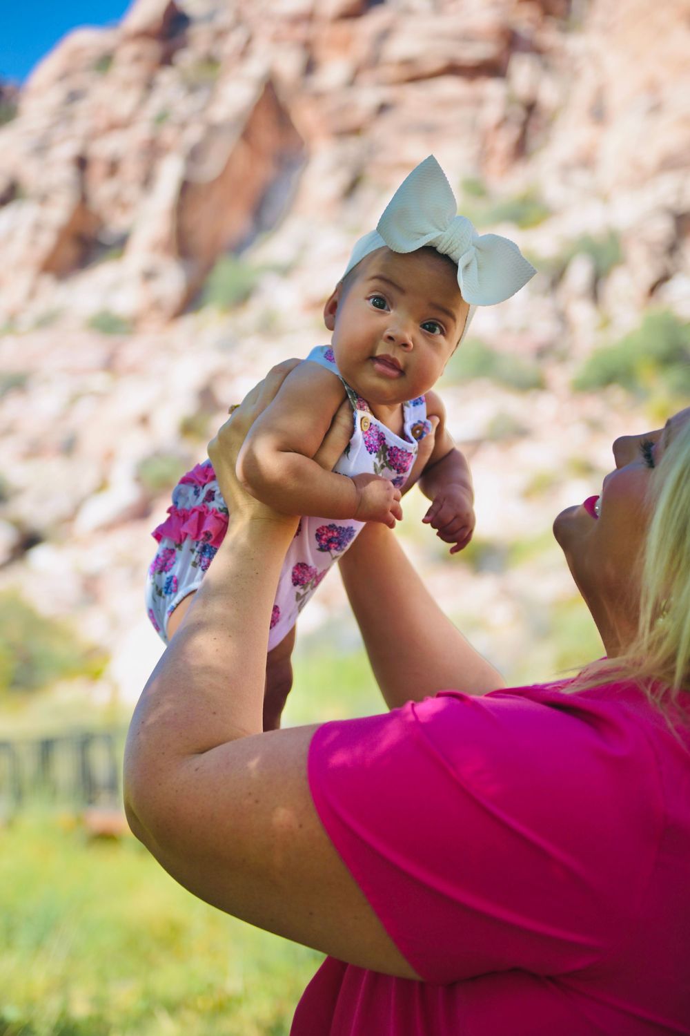 A woman in a pink top lifts a baby girl wearing a white bow and floral dress outdoors near red rock formations.