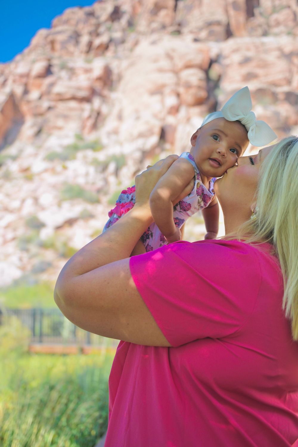 Woman in pink shirt holds baby with white bow outdoors near red rock canyon landscape.