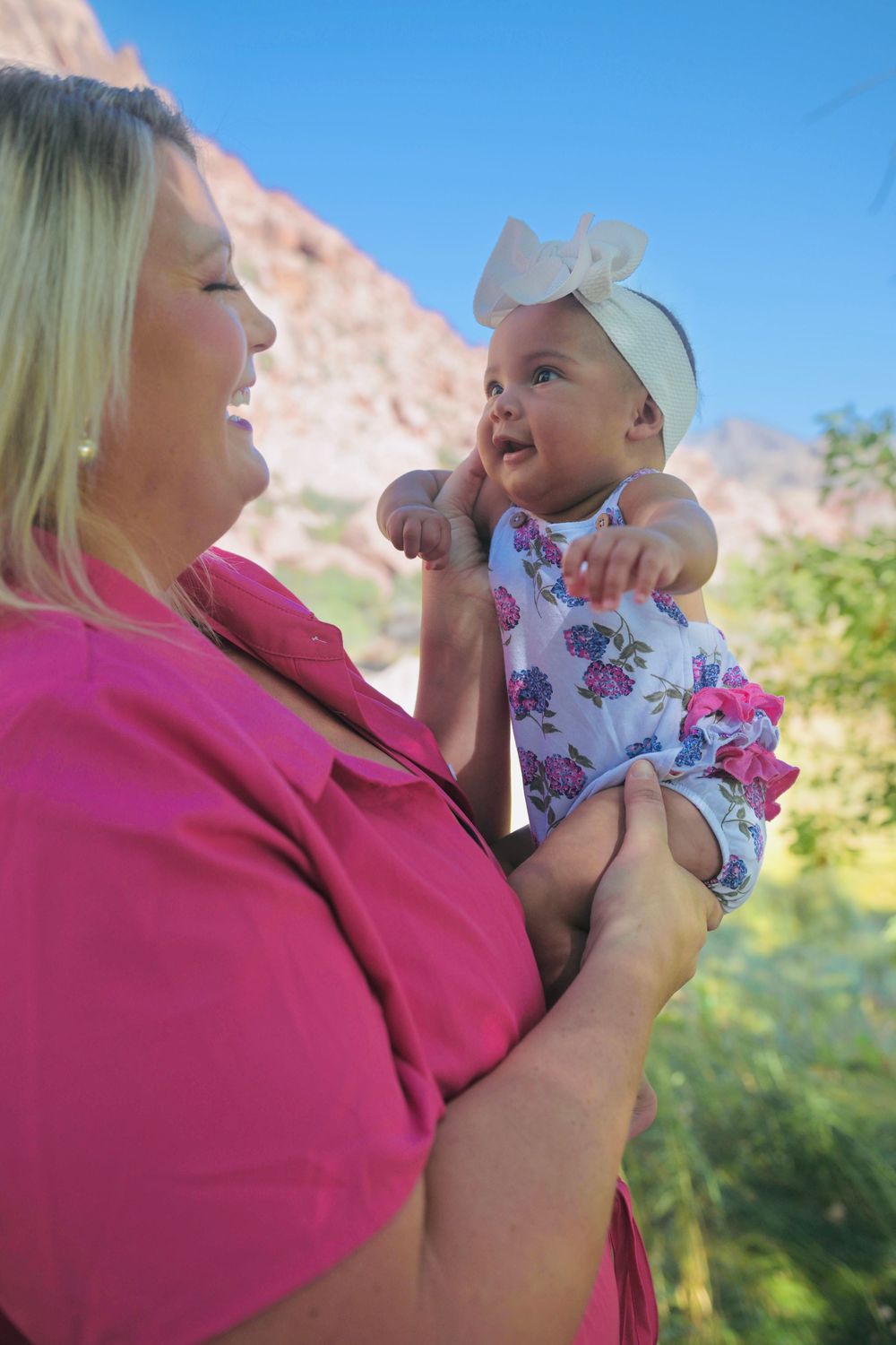 Blonde woman in pink dress lifts a smiling baby girl in a floral outfit near red rocks.