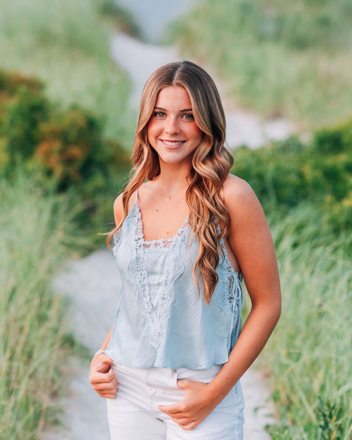 Young woman in light blue lace tank top and white jeans smiling on a sandy path surrounded by greenery.