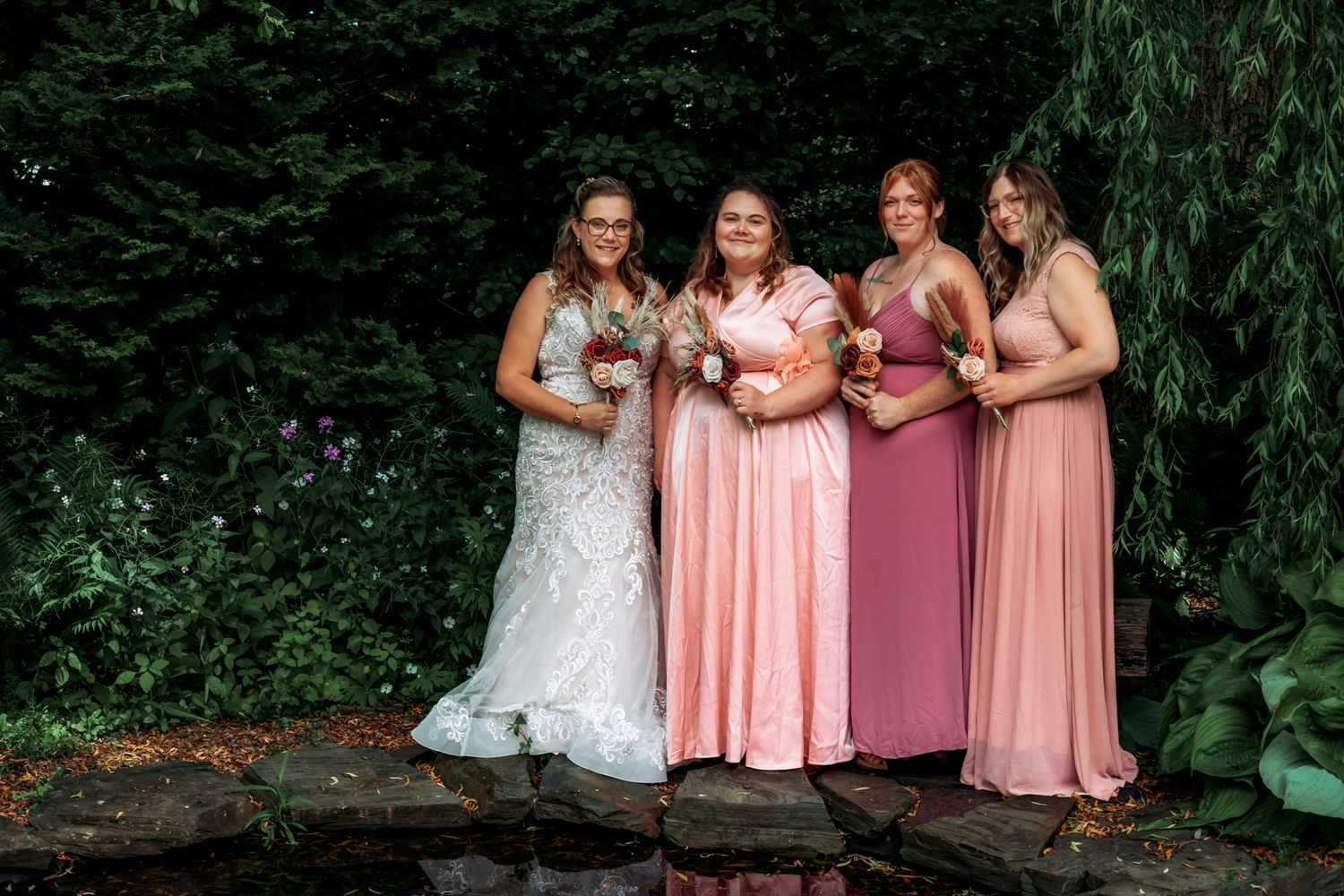 Bride in white lace gown poses with three bridesmaids in pink dresses outdoors near a pond with lush greenery.