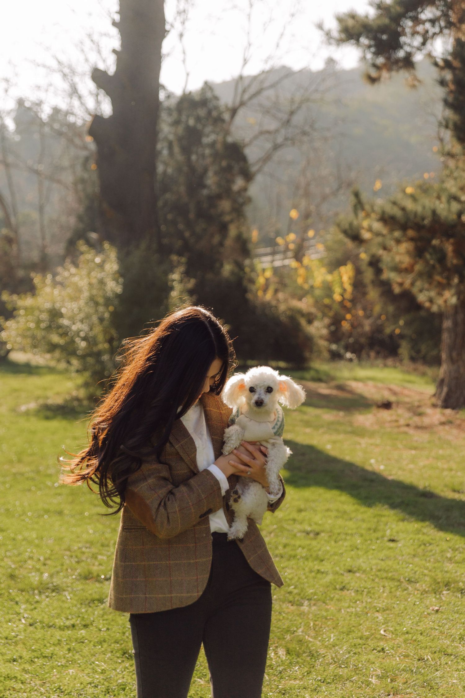Young woman with long dark hair holding a small white fluffy dog in a sunny park.