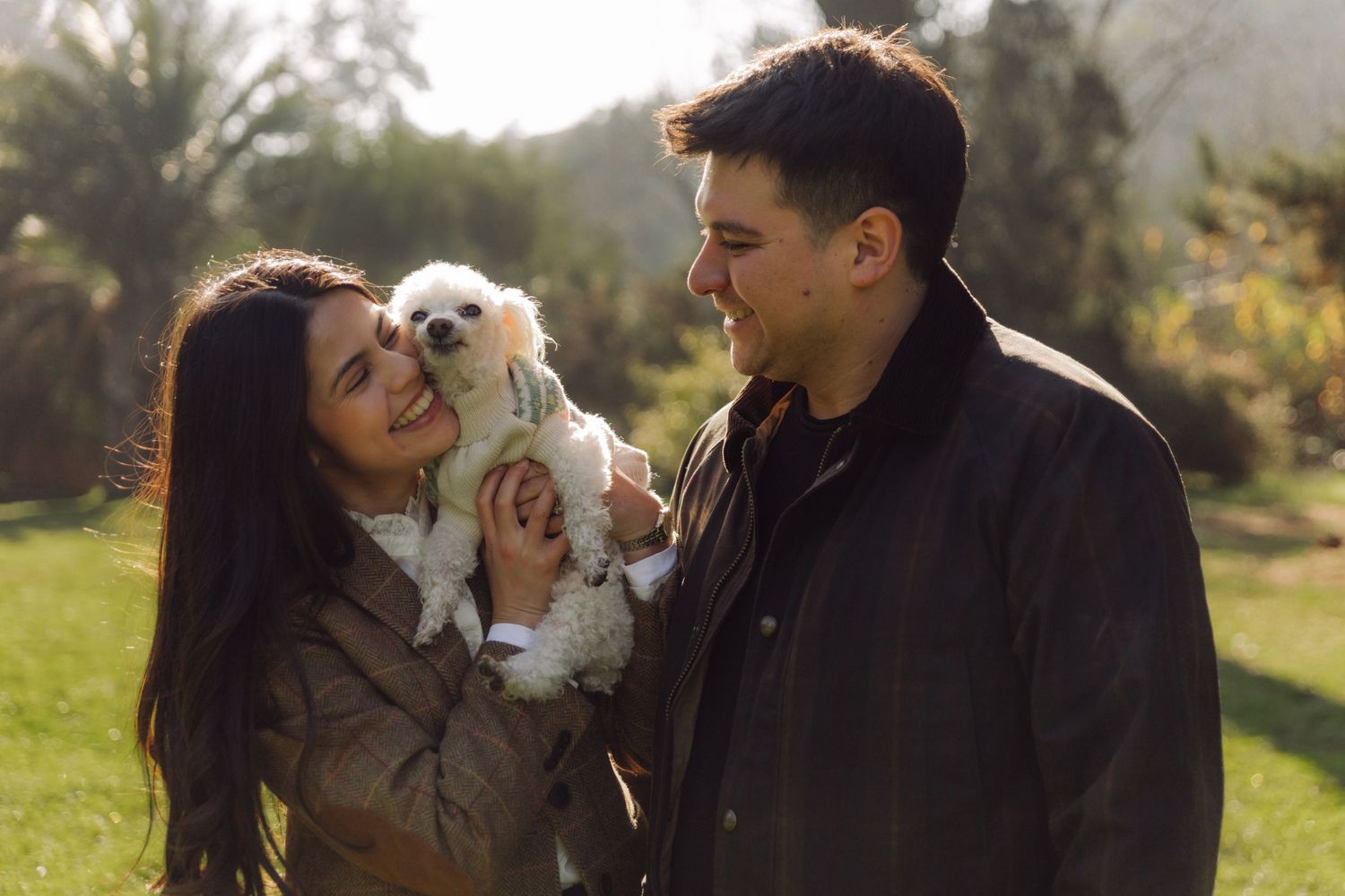Happy couple smiling at each other while holding a small white fluffy dog in a sunny park.