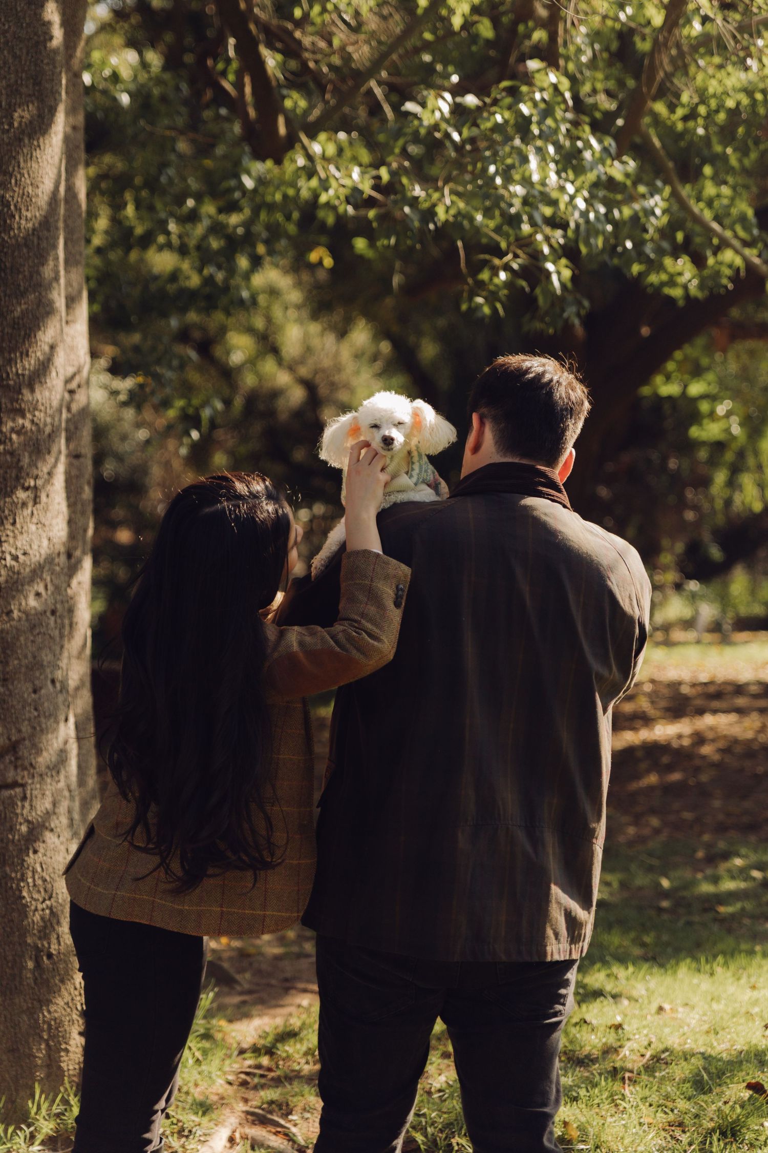Couple from behind holding a small white dog up in a shaded park.