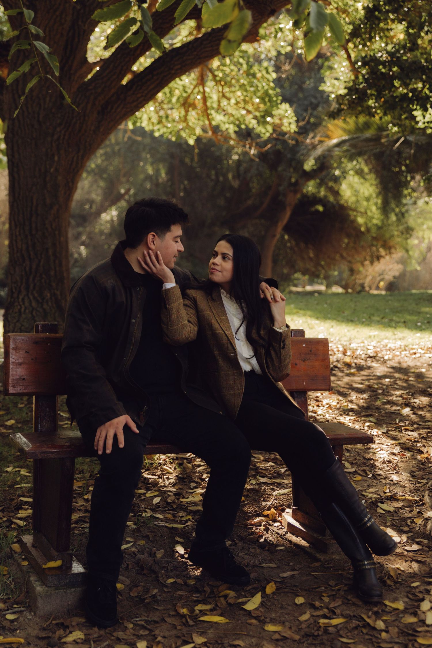 Couple sitting close together on a wooden bench under a large tree in autumn park.