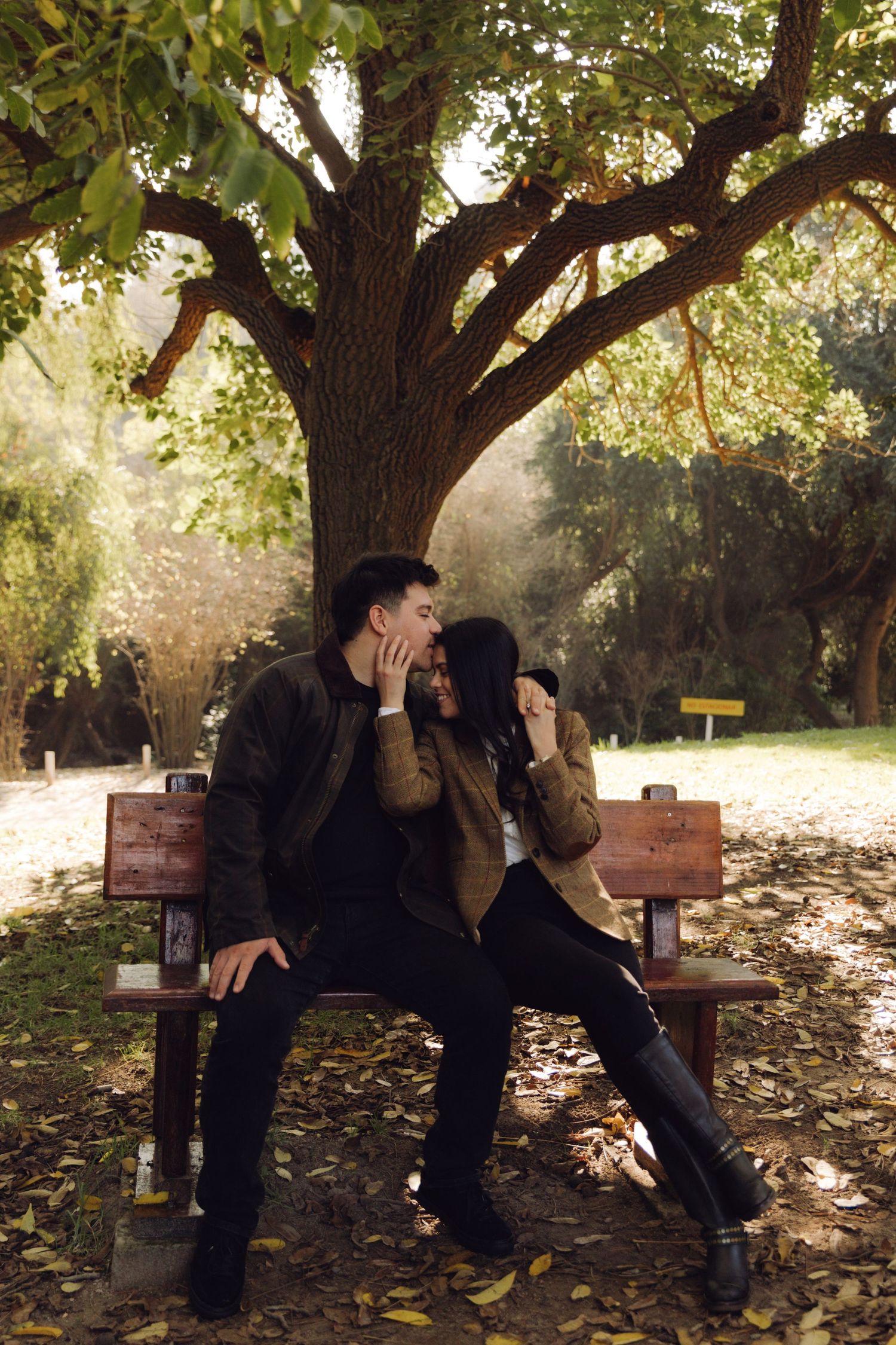 Couple leaning close together on a park bench surrounded by fallen autumn leaves.
