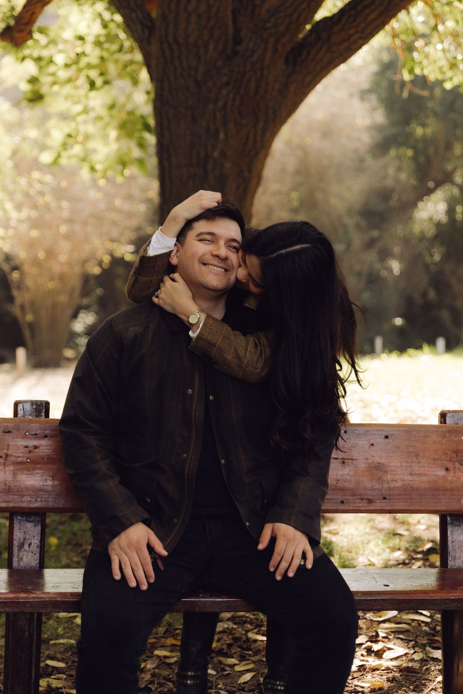Woman kissing smiling mans cheek while sitting on a bench in a sunny park.